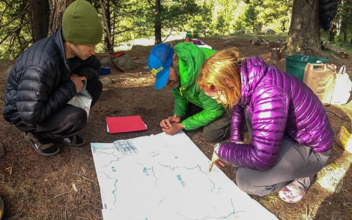 Three people examine a map that is spread out on the ground in a wooded area. 
